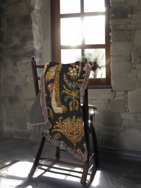 A rectangular, 100% woven cotton throw blanket with a detailed jacquard design. The throw features a symmetrical, vertical pattern of two howling wolves in profile, rendered in shades of beige, gold, and brown against a solid black background. The wolves are surrounded by intricate floral and foliage motifs. The thick, textured weave is visible. The blanket is finished on all four sides with a two-inch, multi-colored fringe that includes black, white, red, and gold threads.